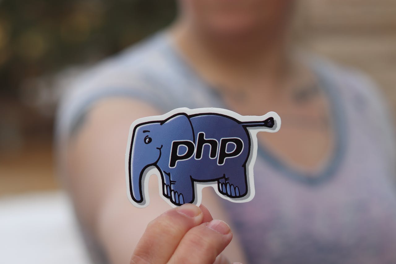 A woman's hand holding a PHP elephant mascot sticker, blurred background.